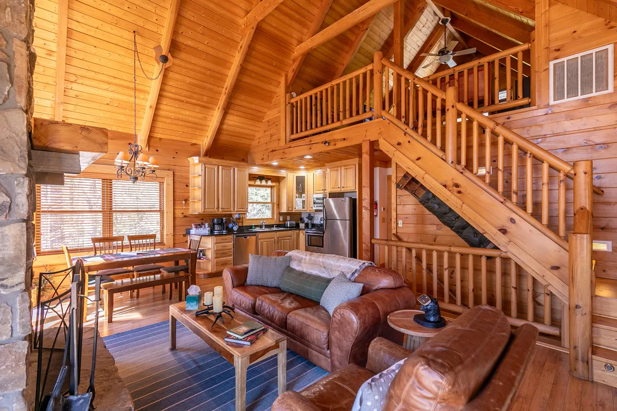 A wide-angle view highlighting the open flow among the living room, kitchen, and dining area. The image features a comfortable sofa facing the stone fireplace, a modern kitchen with stainless steel appliances, granite countertops, and a staircase leading to other parts of the cabin.