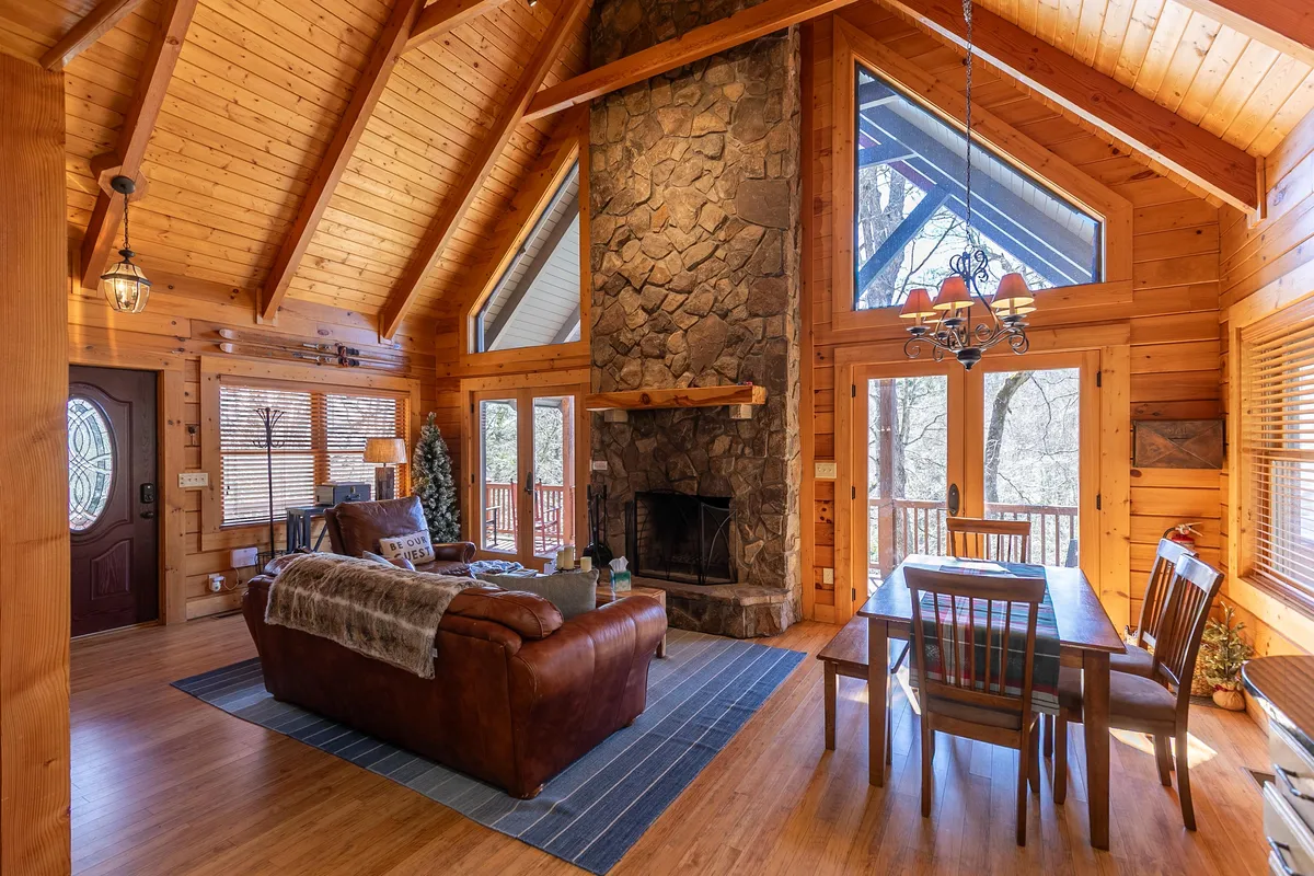 A warm and welcoming open-concept living room with a grand stone fireplace as its centerpiece. Large windows reveal sweeping forest views, while exposed wooden beams add rustic character. A plush leather sofa and a spacious dining table create an inviting space for gatherings, relaxation, and meals with family and friends.