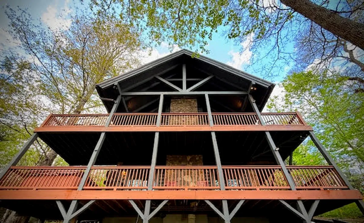 The image showcases the exterior of the cabin, with prominent wooden balconies overlooking lush, natural surroundings. The setting sun casts a warm glow, creating an inviting and serene mountain retreat atmosphere. The scene suggests opportunities for relaxing on the balcony, enjoying nearby nature walks, and gathering with family.