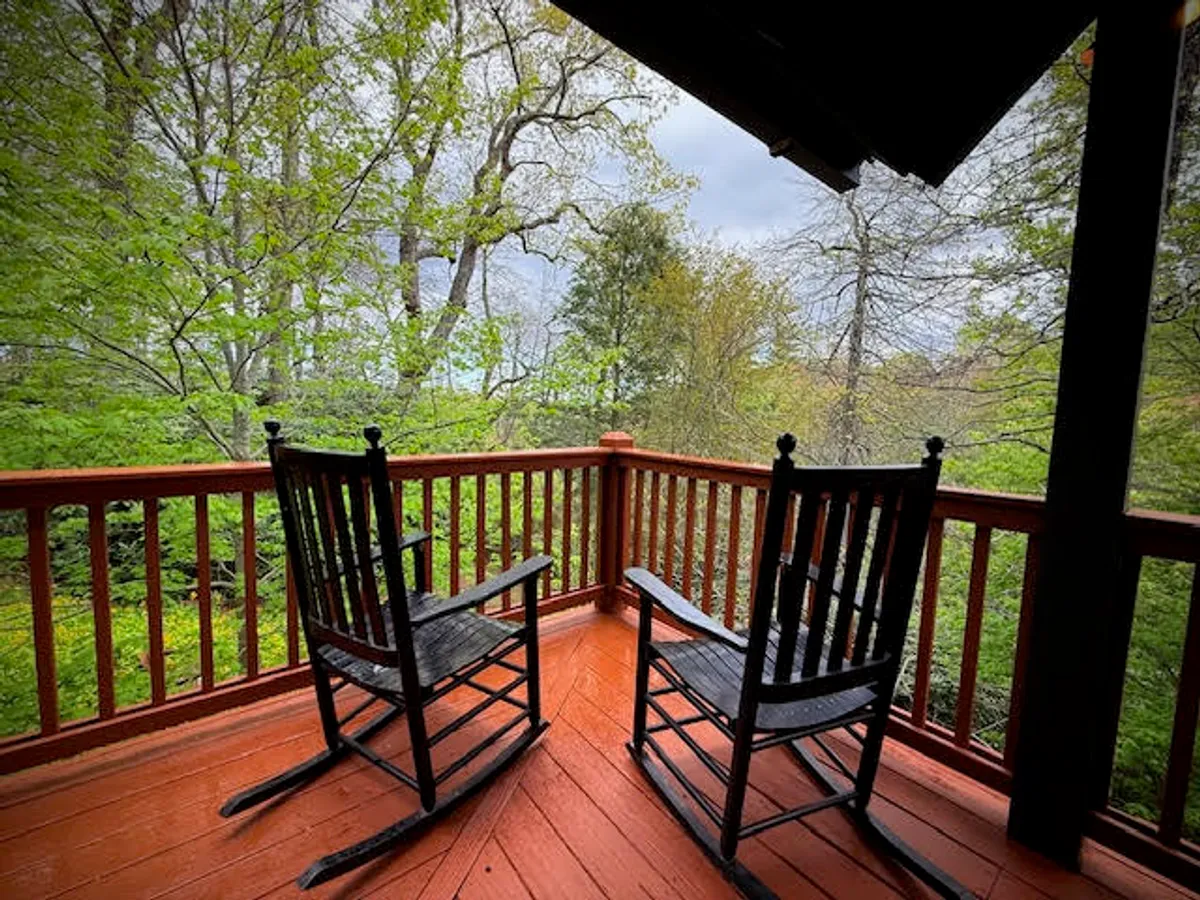 A charming exterior porch scene with classic wooden rocking chairs set along a sturdy wooden railing, overlooking vibrant greenery. The soft morning or evening light creates a tranquil atmosphere, inviting guests to relax and soak in the natural surroundings. The scene suggests a spot perfect for watching sunrises, sunsets, or stargazing after a day of adventure.
