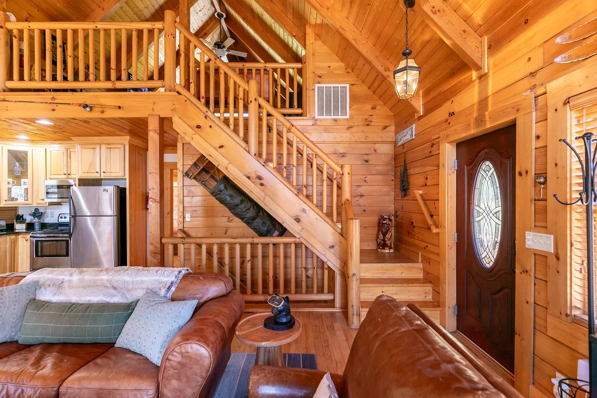 This image captures the main entrance of the cabin, with a view of the wooden staircase, a leather sofa, the kitchen area, and a prominent front door. Natural light filters in, creating a welcoming and warm environment that feels connected to the outdoors.