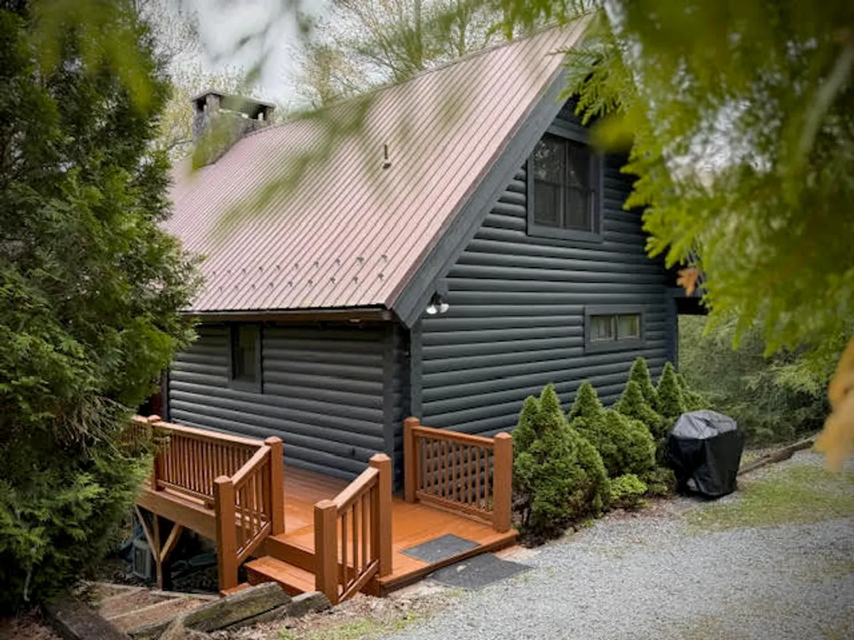 This image showcases the inviting exterior of a charming log cabin nestled among lush green foliage, with a spacious wooden deck perfect for gatherings. A gravel path leads up to the cabin, highlighting its secluded forest location and tranquil ambiance.