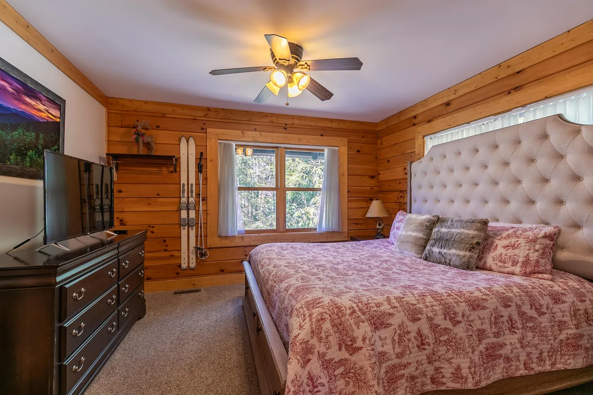 A serene bedroom with a tufted headboard, large window framing forest views, TV stand with television, and ski equipment decorating the wall. The plush bedding and natural light create a restful, inviting ambiance.