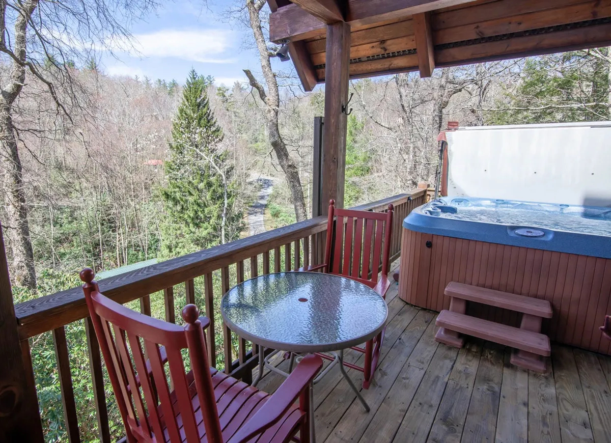 An exterior shot highlighting a tranquil deck area featuring a bubbling hot tub, wooden rocking chairs, and a table for socializing. The lush trees surrounding the deck emphasize privacy and immersion in nature. The mood is tranquil and inviting, ideal for unwinding after a day of outdoor activities.