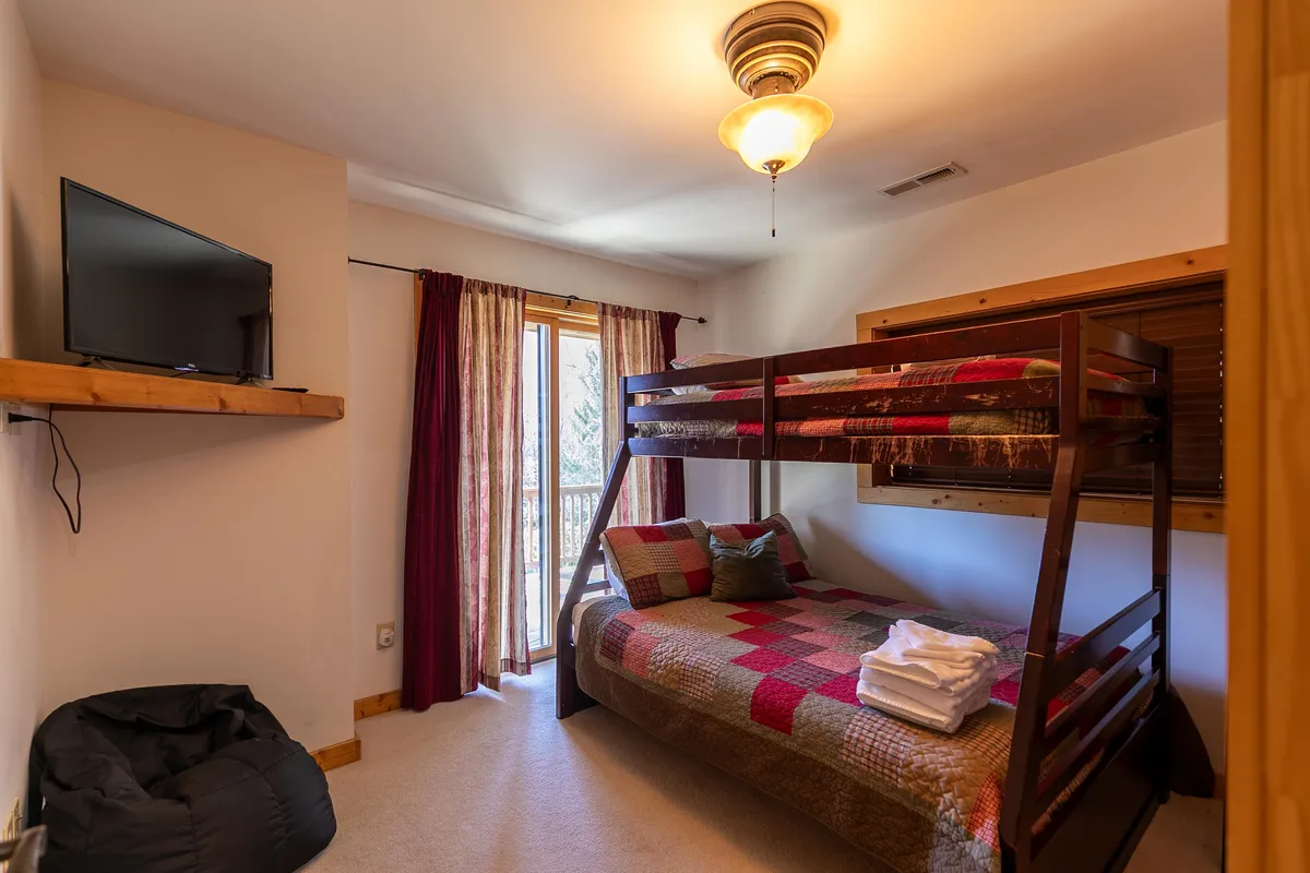 A charming bunk room with a plaid-patterned bunk bed, window with curtains letting in soft light, and a television. The space feels inviting and perfect for families or kids, with a warm, cozy atmosphere.