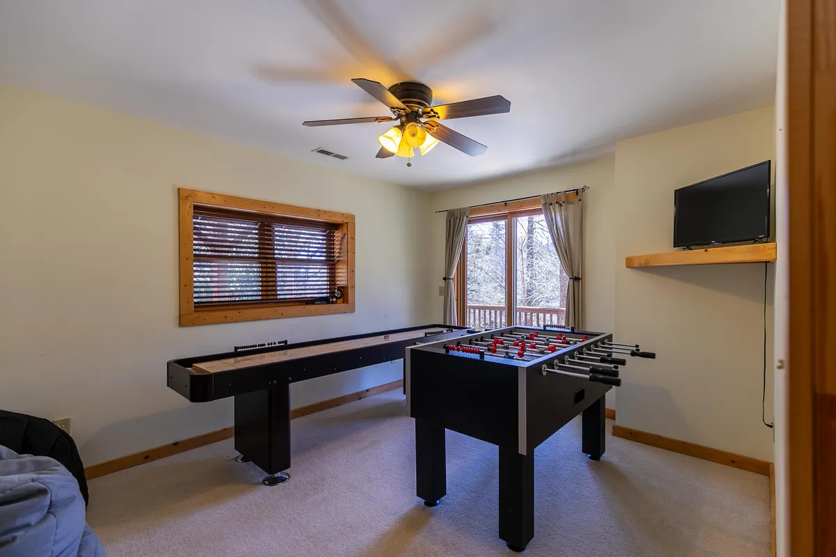 This image features a vibrant game room with both a foosball table and shuffleboard table, a large window providing natural light, and space for socializing. The setup suggests an inviting space for spirited games and casual relaxation.