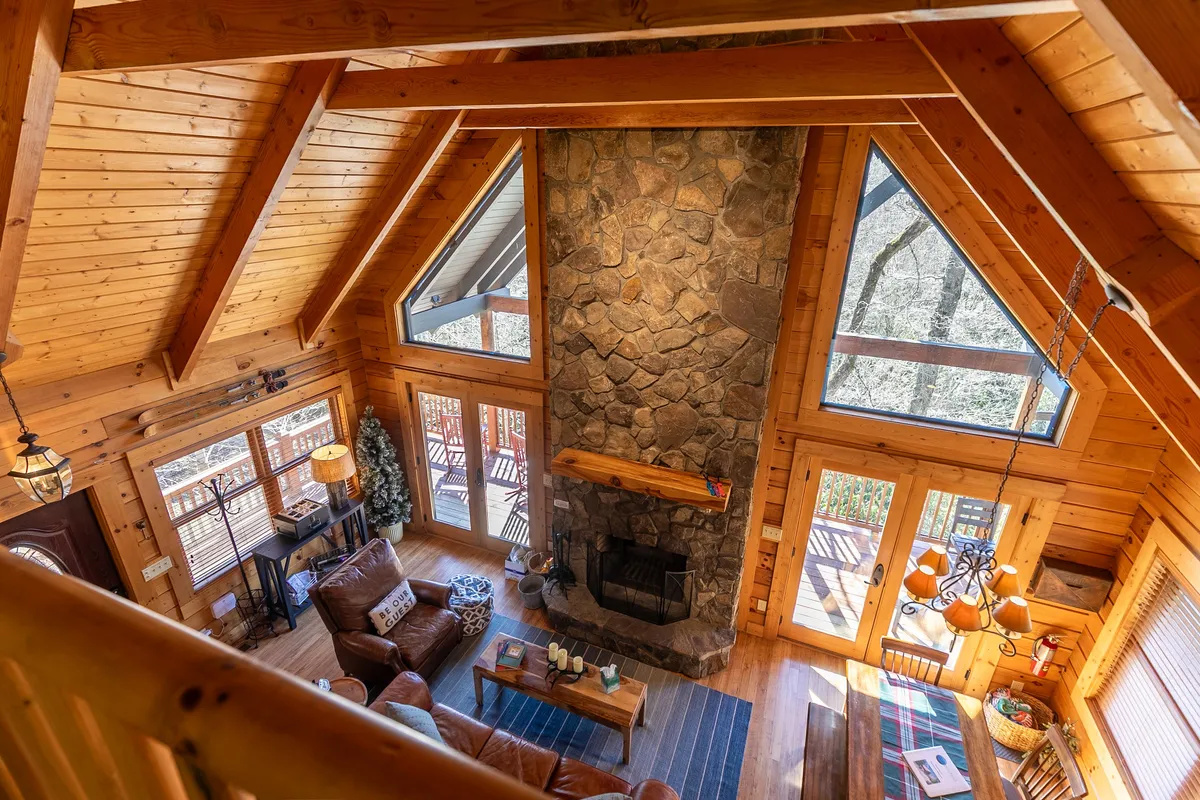 A spacious living area featuring a striking stone fireplace, large windows framing the natural surroundings, exposed wooden beams, and an inviting seating area. The view through the windows hints at the scenic setting outside, tying the interior comfort to the beautiful environment the cabin is known for.