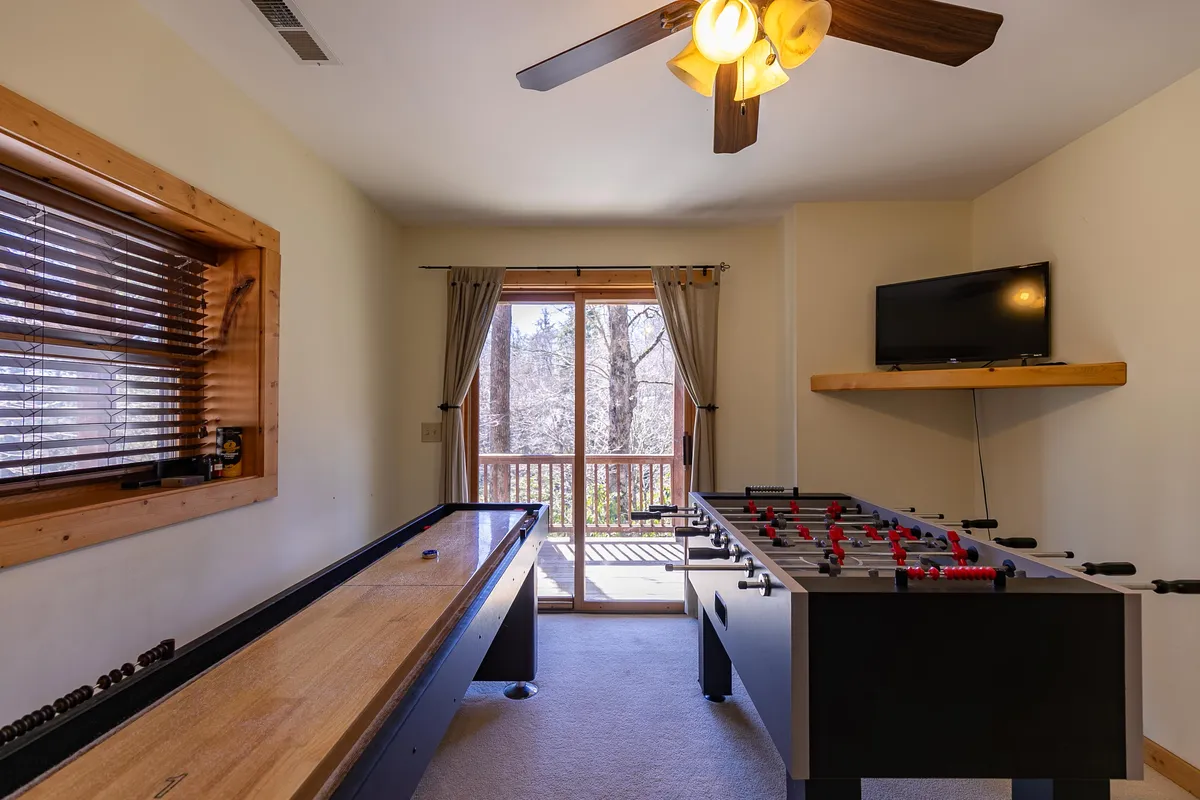 A vibrant interior scene of the cabin's game room, featuring a shuffleboard table, foosball table, TV, and a large window that connects the indoors to the scenic outdoors. The setup invites friendly family competitions and relaxed socializing, with views of the natural environment beyond.