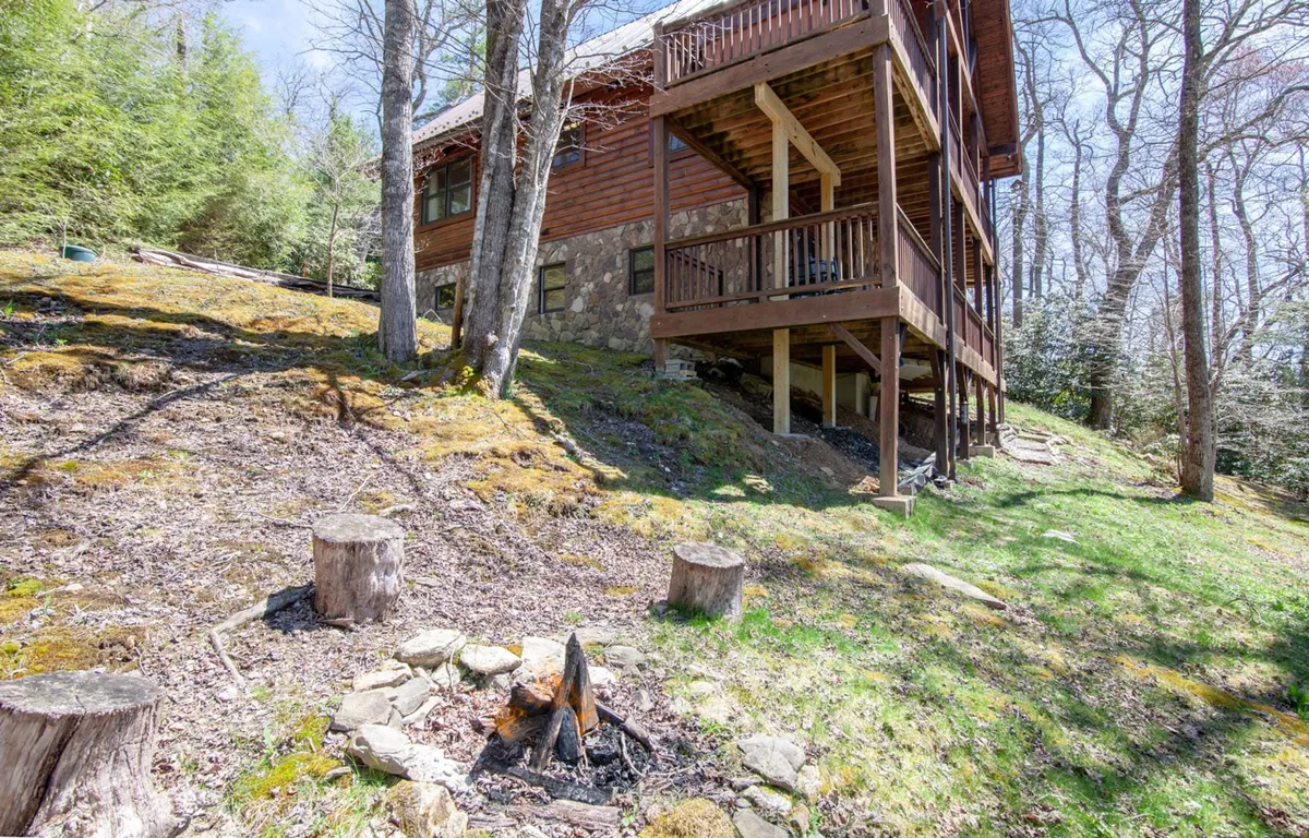 A charming wooden house with a prominent fire pit area in the foreground, set amid tall trees. The fire pit, encircled by seating, is perfectly positioned for evening gatherings, storytelling, and stargazing. The natural setting enhances the mood of cozy outdoor adventure.
