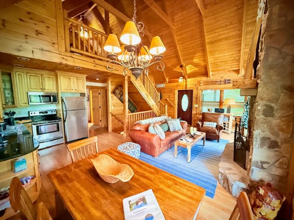 An inviting open-concept living and dining area featuring exposed wooden beams, a striking stone fireplace, plush sofas with cushions, and a large dining table set for gatherings. The warm, welcoming atmosphere is enhanced by soft lighting, suggesting a space where groups can congregate for meals, games, or fireside chats.