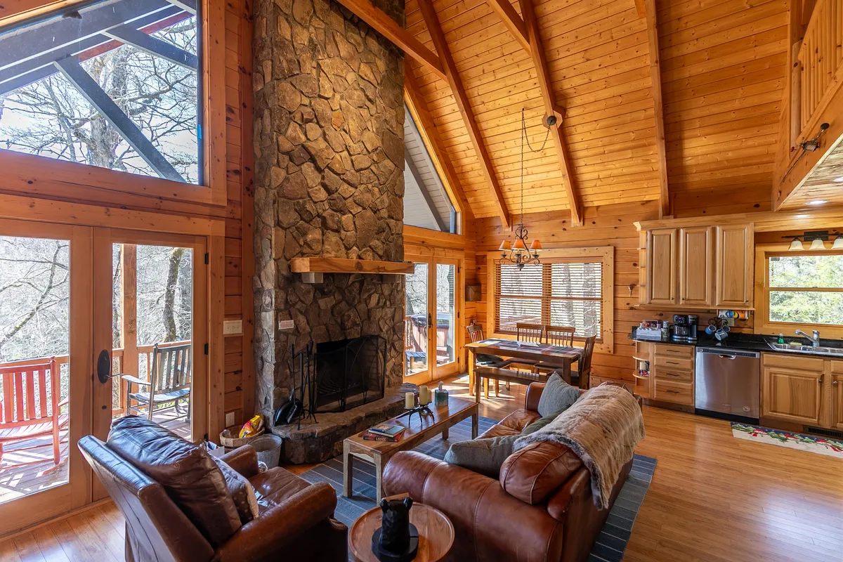 An inviting living and dining area featuring a stone fireplace, expansive windows with views of the surrounding woods, substantial wooden beams, and a rustic-chic dining table. The scene radiates warmth and comfort, ideal for sipping coffee at sunrise or gathering for a meal.