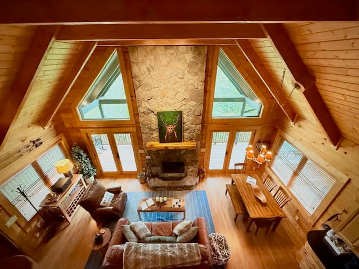 A spacious open living area featuring a stone fireplace, large windows with views of the outside, wooden beams, a dining table, and a comfortable sofa. The expansive windows visually connect the cozy interior to the surrounding Blue Ridge Mountain landscape.